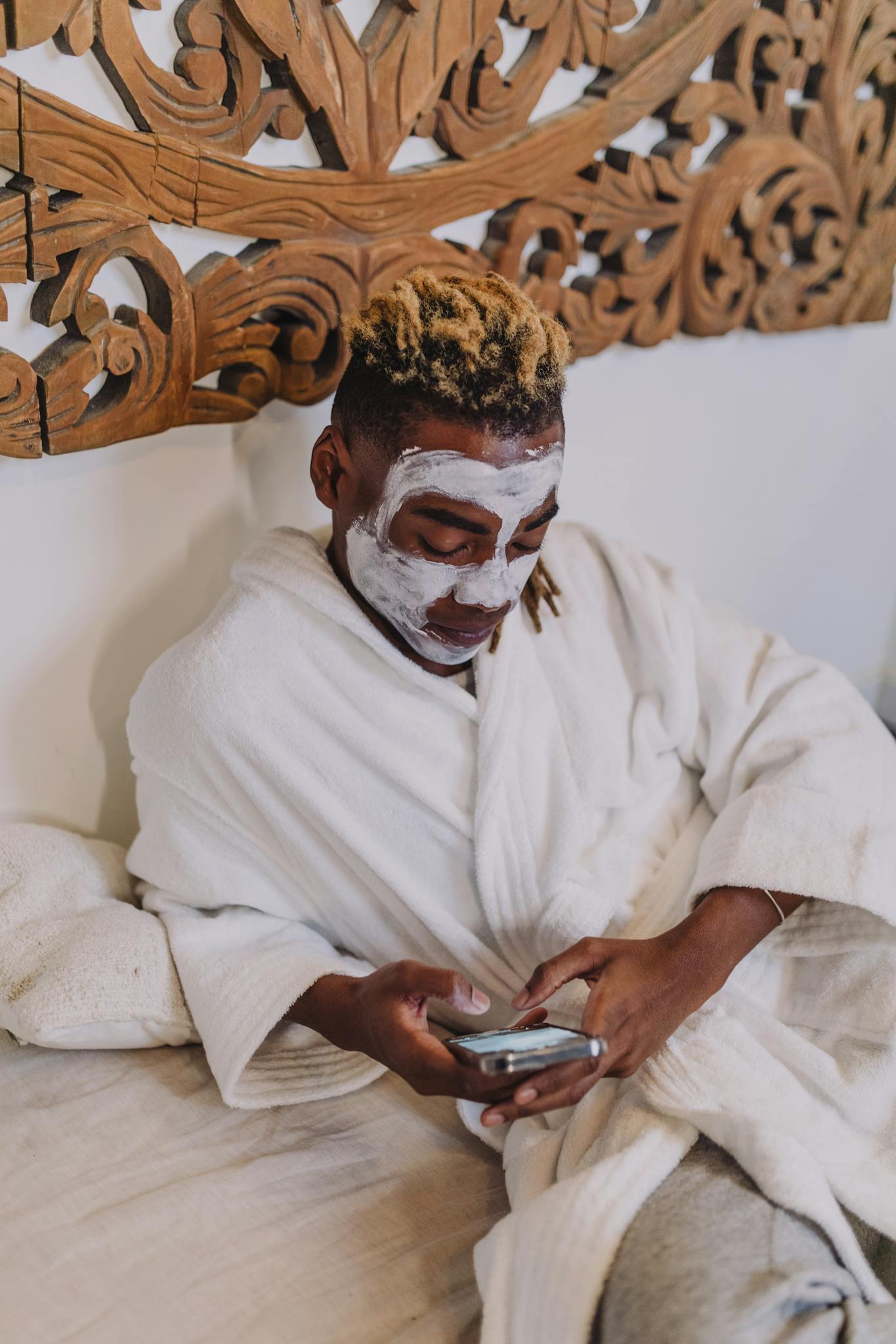 A person enjoying a spa day with a facial mask and using a phone, indoors.