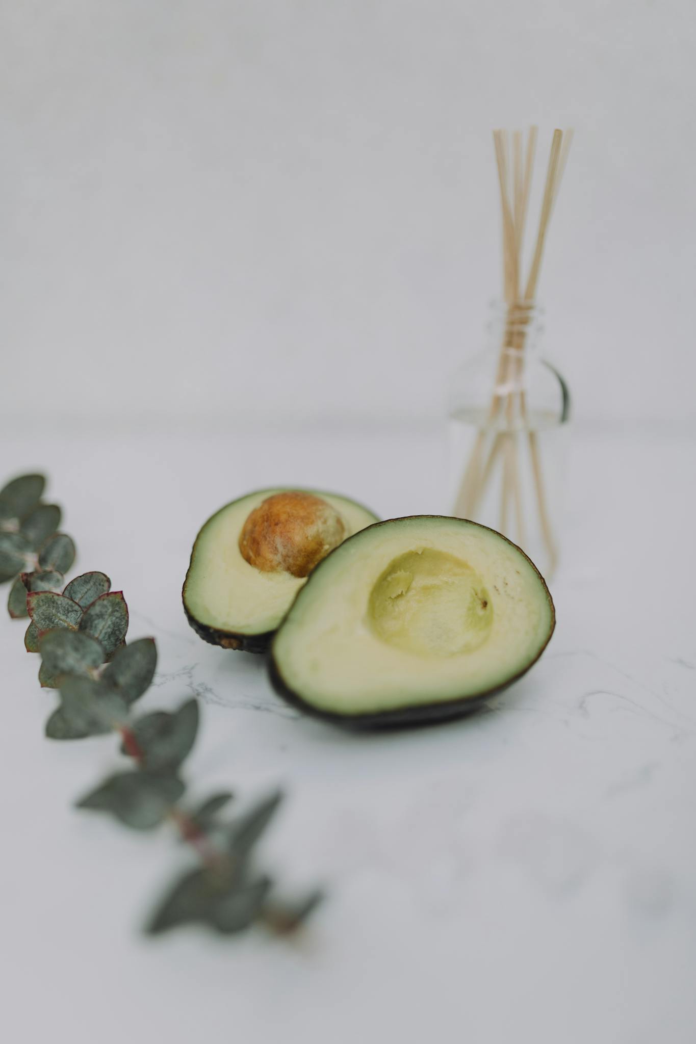 A serene still life featuring sliced avocado with eucalyptus leaves and a reed diffuser.
